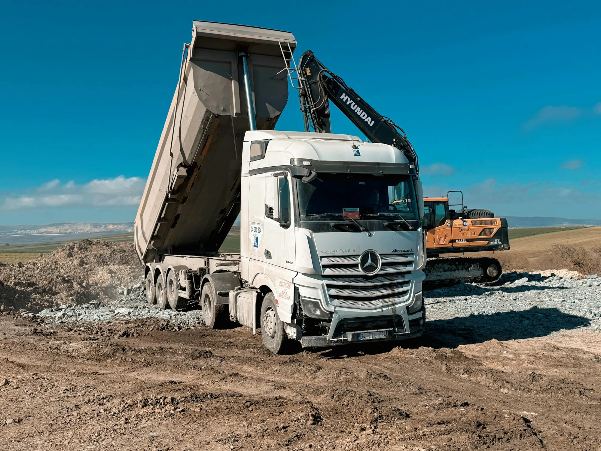 Camión volquete de servicio pesado y excavadora operando en un día soleado en un sitio de construcción.