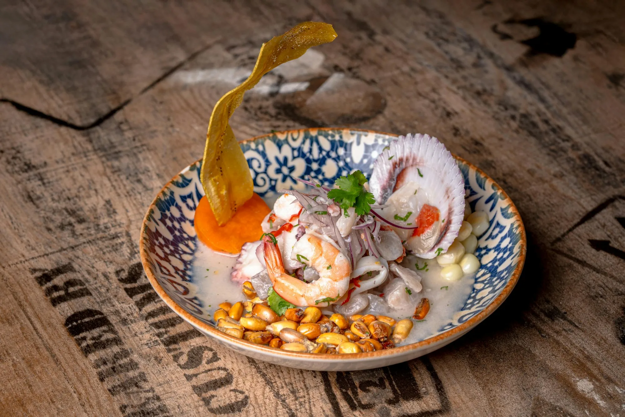 Primer plano de un ceviche tradicional de mariscos con camarones, almejas y chips de plátano en una mesa de madera rústica.