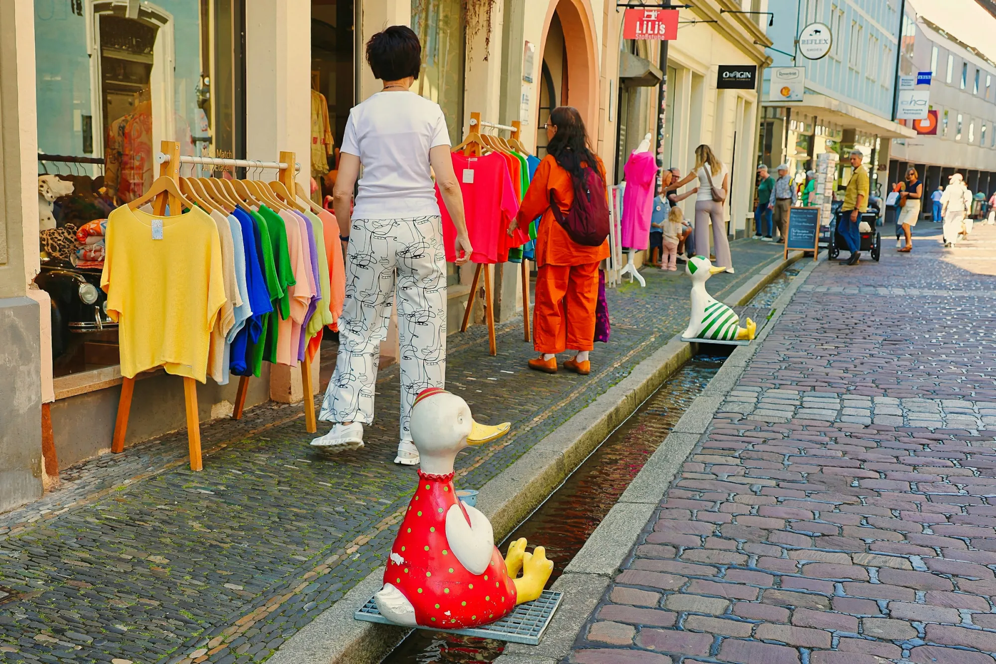 People walk past storefronts and decorative ducks along a street.