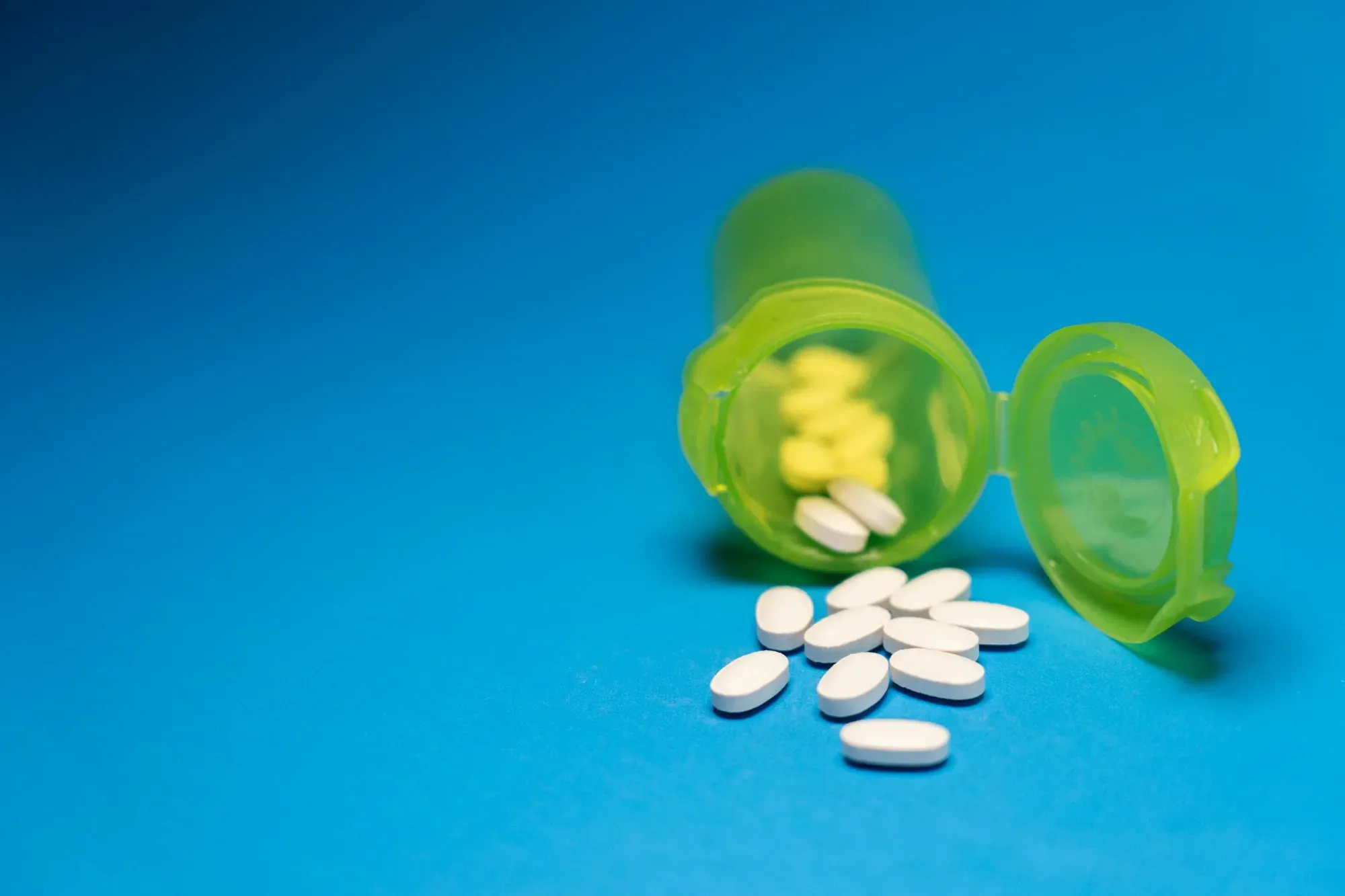 Imagen de un frasco de pastillas verde con pastillas blancas esparcidas sobre un fondo azul, que simboliza la atención médica y la medicación.